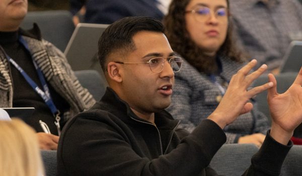 A student sitting in a lecture hall speaks, gesturing with both hands, as other students look on
