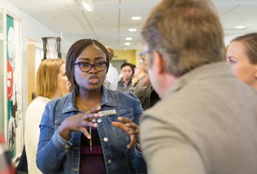 A Black woman wearing a denim jacket and glasses explains a concept to a man at a poster session.