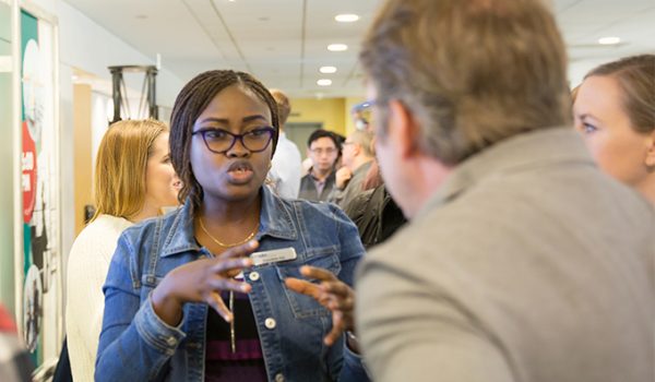 A Black woman wearing a denim jacket and glasses explains a concept to a man at a poster session.
