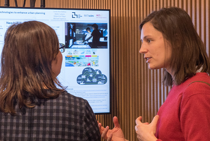 A woman in a red sweater explains a concept to another woman as they stand in front of a monitor displaying a poster