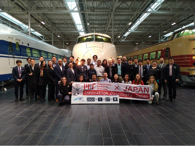 A group of students and staff stand in front of decommissioned trains, holding a banner with the MIT logo and "Innovation Discovery Japan".