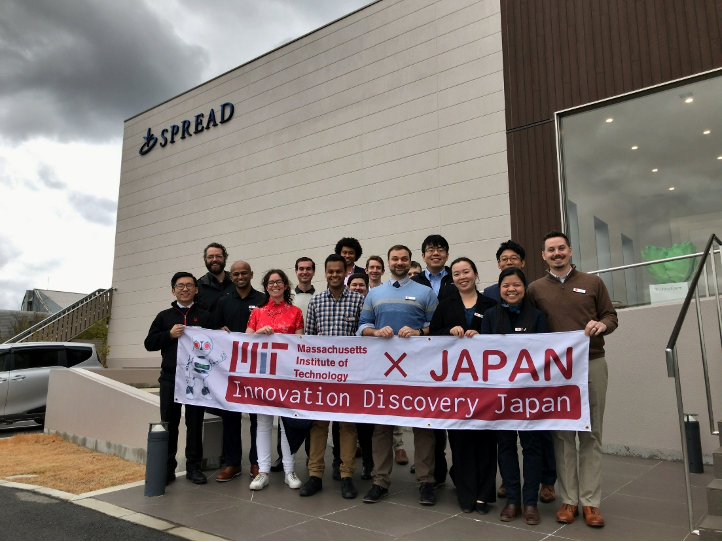 A group of students holding the MIT IDJ banner stands outside the Spread building, with the company's logo on the exterior wall.