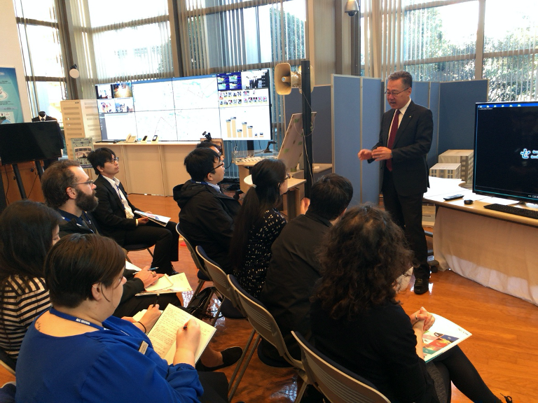 Yuji Misaki stands before a group of seated students in a large room with several displays