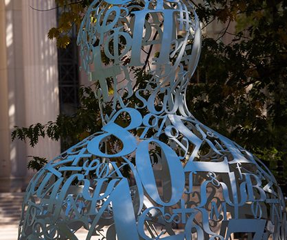 The Alchemist, a sculpture made up of large curved numbers and symbols in the shape of the head and shoulders of a person, is in the foreground. In the background are the pillars and steps of the main entrance to MIT.