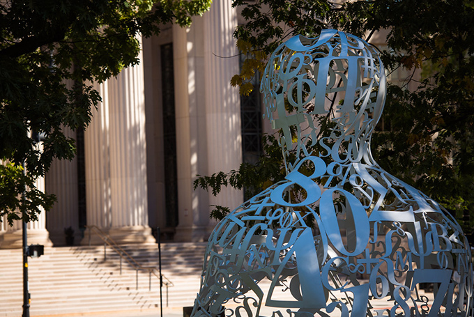 The Alchemist, a sculpture made up of large curved numbers and symbols in the shape of the head and shoulders of a person, is in the foreground. In the background are the pillars and steps of the main entrance to MIT.