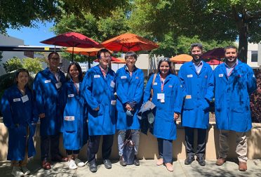 A group of students in long blue lab coats, standing outside in partial sun and partial shadow.