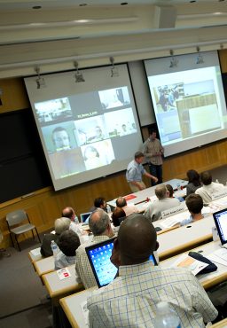 A lecture hall with two projector screens down at the front of the room, one screen filled with images of people who are participating in a video conference.