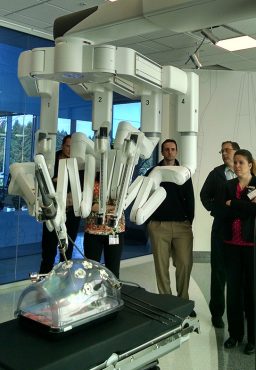 Students stand around an elaborate surgical robot, which has multiple segmented arms suspended from a central structure in midair.