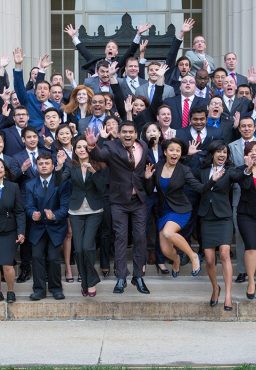 A large group of students stand on stone steps. They are all in formal business attire and many of them are making silly faces or jumping in the air.