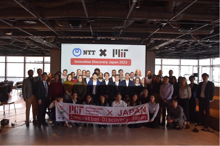 Students and NTT staff stand in a group holding an MIT x Japan banner.