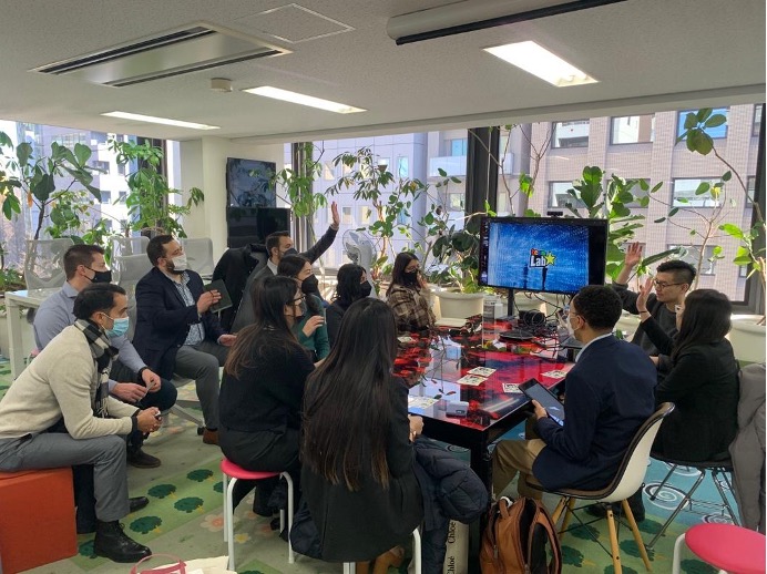 Students and teamLab staff gather around a table in an office with plants all around the edges of the room.