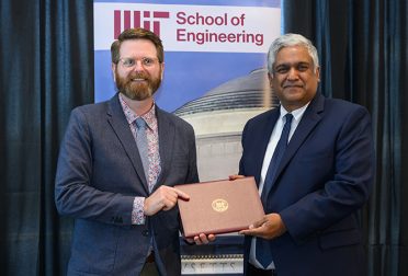 Steven Derocher, a tall white man with red hair, a beard, and glasses, receives an award from Anantha Chandrakasan, a slightly shorter man with brown skin and white hair. They are standing in front of a banner for the MIT School of Engineering.