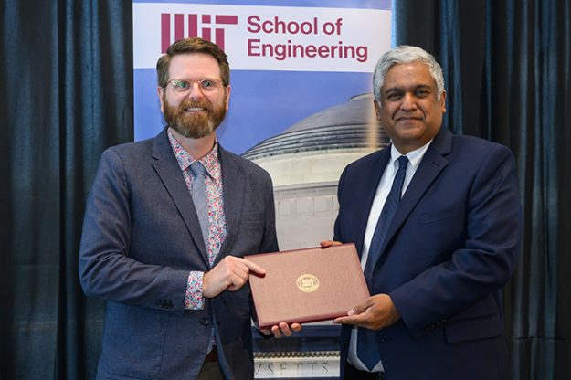 Steven Derocher, a tall white man with red hair, a beard, and glasses, receives an award from Anantha Chandrakasan, a slightly shorter man with brown skin and white hair. They are standing in front of a banner for the MIT School of Engineering.