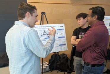 Three male students gather around a poster on an easel. One is gesturing in the air, a second is listening intently, and the third is laughing.