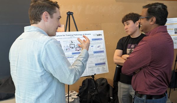 Three male students gather around a poster on an easel. One is gesturing in the air, a second is listening intently, and the third is laughing.