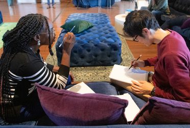 Two students sit on a couch together, with one explaining something and gesturing and the other drawing on a piece of paper.