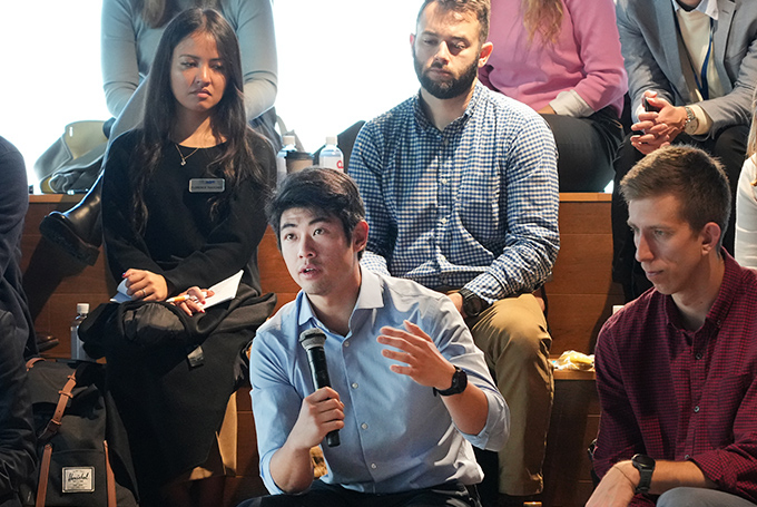 A group of students sitting on wooden risers. One holds a microphone and is asking a question, gesturing with his free hand.