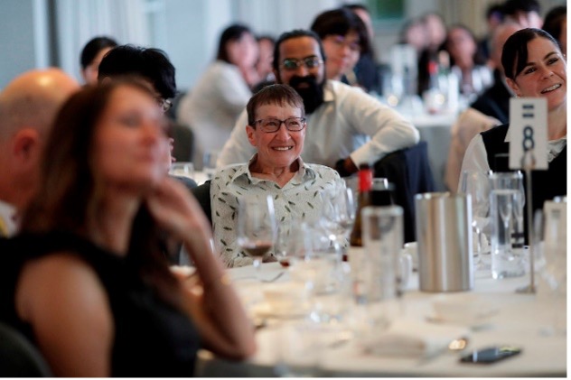 Linda Newnes sits at a banquet table, smiling. There are other people in the picture but they are out of focus.