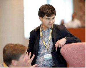 A man standing and leaning against the back of a chair, listening to a conversation.