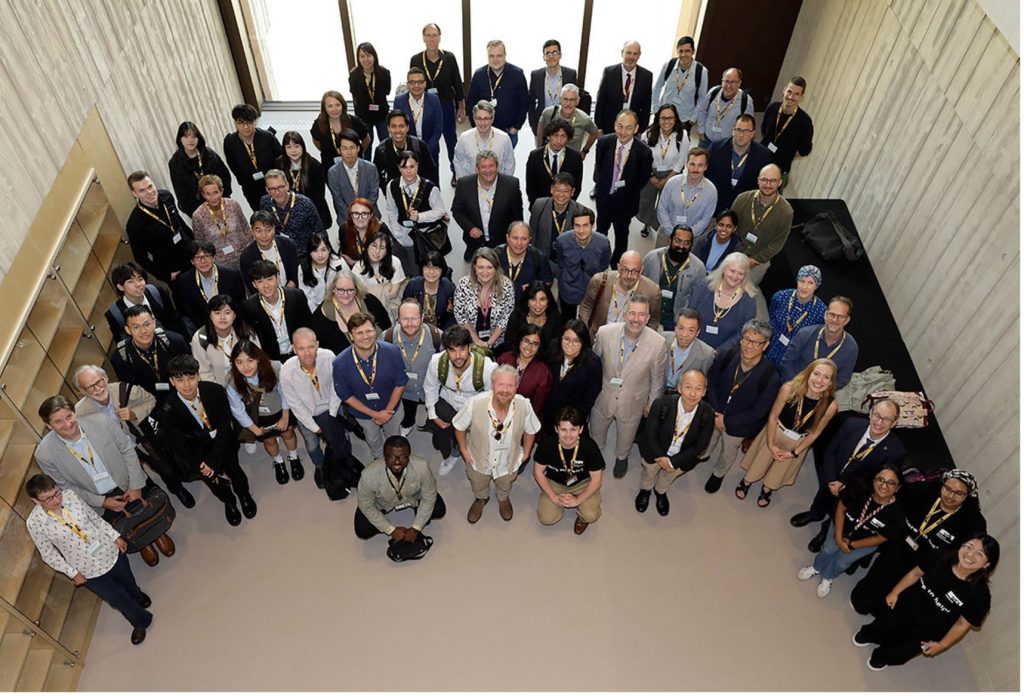 A large group of people in a building atrium, photographed from above. Everyone is looking up at the photographer. 