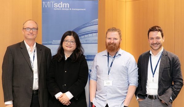 John Hajdukiewicz, Charlotte Wang, Doug Hamilton, and Nick Borge stand together in front of a pop-up banner with the SDM logo. They all smile for the camera.