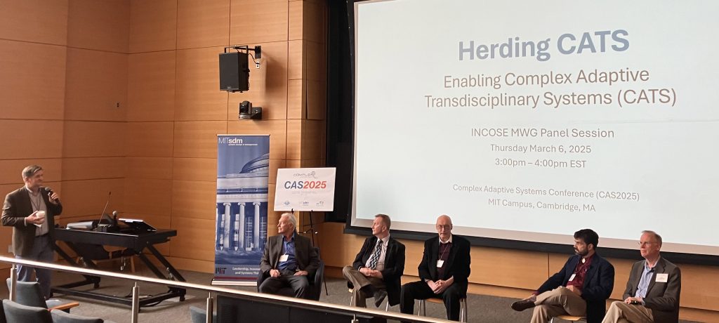 A group of five men sitting in chairs at the front of a lecture auditorium, with a sixth man standing to the left with a microphone. Behind the sitting men is a projection screen that reads "Herding CATS: Enabling Complex Adaptive Transdisciplinary Systems (CATS)"