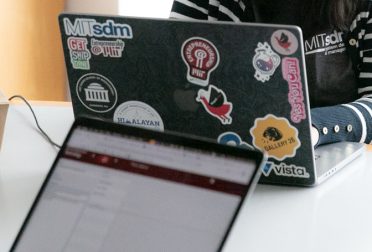 Two laptop computers, one with the screen facing the camera and one with the case facing the camera. The laptop case facing the camera has a number of stickers on it including the mitSDM logo and the MIT dome.