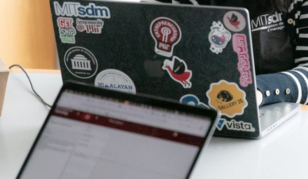 Two laptop computers, one with the screen facing the camera and one with the case facing the camera. The laptop case facing the camera has a number of stickers on it including the mitSDM logo and the MIT dome.