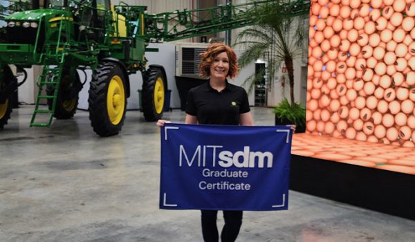 Rebecca Halverson, holding an MITSDM Graduate certificate flag, standing in front of a large piece of farm machinery and a screen showing an image of beans.
