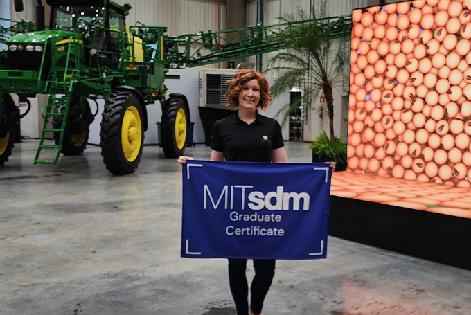 Rebecca Halverson, holding an MITSDM Graduate certificate flag, standing in front of a large piece of farm machinery and a screen showing an image of beans.