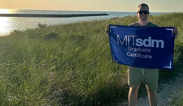 John Hushaw holding a flag reading "MITsdm Graduate Certificate," standing among tall grass with water in the background.
