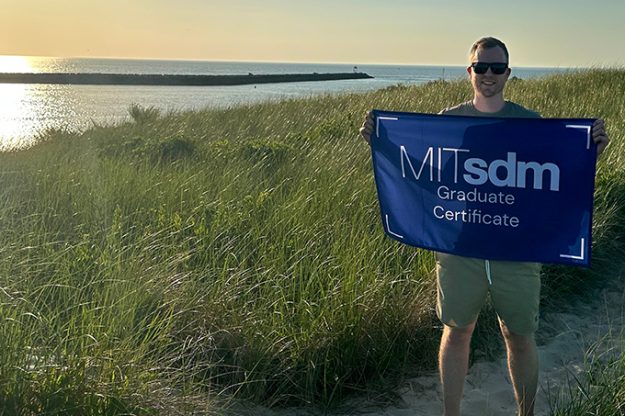 John Hushaw holding a flag reading "MITsdm Graduate Certificate," standing among tall grass with water in the background.