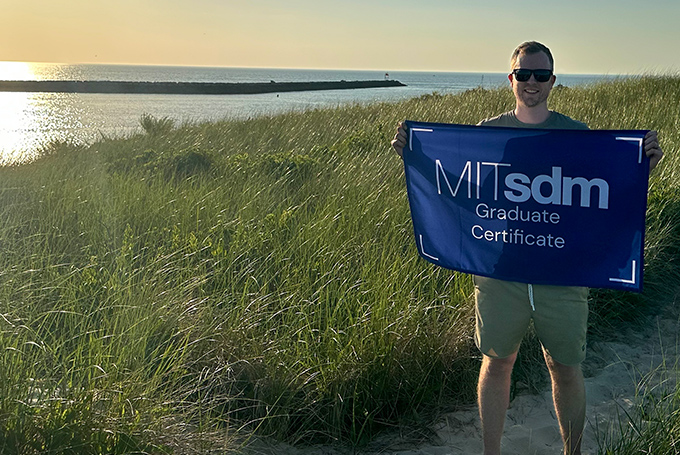 John Hushaw holding a flag reading "MITsdm Graduate Certificate," standing among tall grass with water in the background.