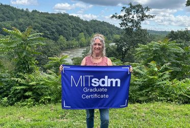Bonnie Novak standing before a lush green landscape with a river behind her. She's holding a blue flag that reads "MITsdm Graduate Certificate."