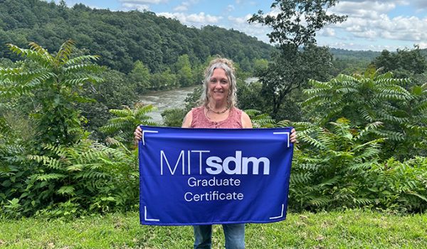 Bonnie Novak standing before a lush green landscape with a river behind her. She's holding a blue flag that reads "MITsdm Graduate Certificate."