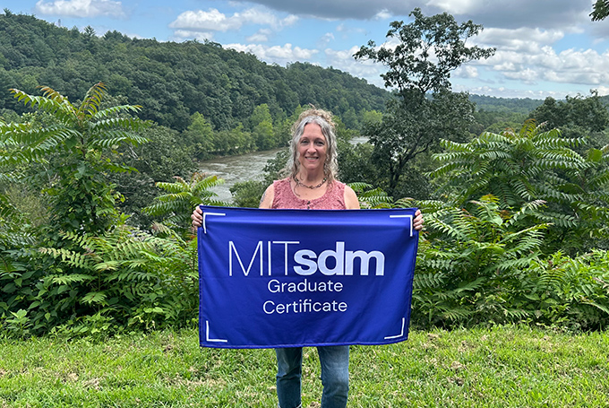 Bonnie Novak standing before a lush green landscape with a river behind her. She's holding a blue flag that reads "MITsdm Graduate Certificate."