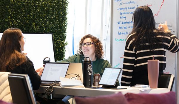 Two students sit at a table smiling while a third stands at a whiteboard, taking notes.