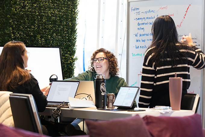 Two students sit at a table smiling while a third stands at a whiteboard, taking notes.
