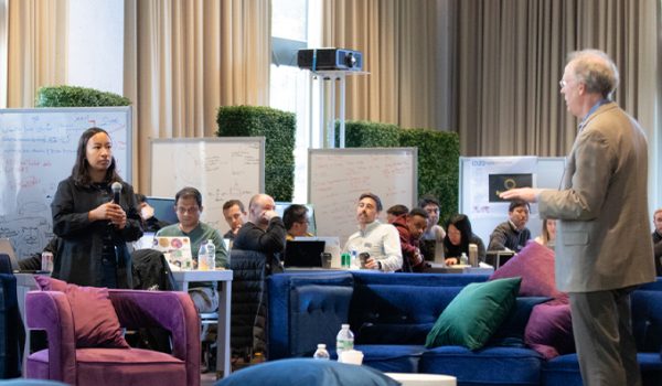 A student stands across from a professor asking a question in a large room with lounge furniture and whiteboards set up around the edges.