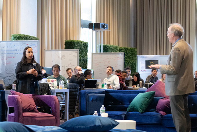 A student stands across from a professor asking a question in a large room with lounge furniture and whiteboards set up around the edges.