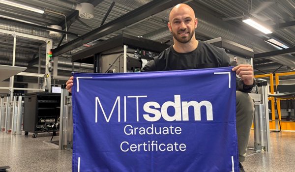 Luc Vernery holding an MITsdm Graduate Certificate banner, kneeling in a warehouse