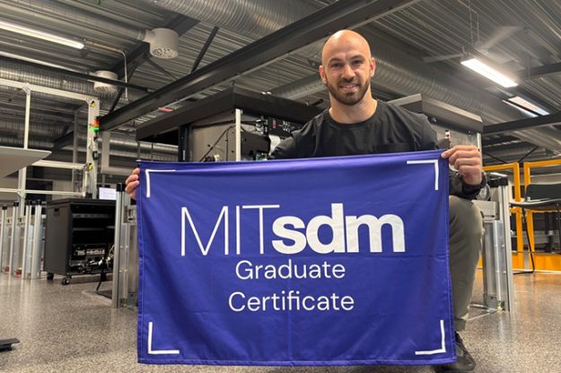 Luc Vernery holding an MITsdm Graduate Certificate banner, kneeling in a warehouse