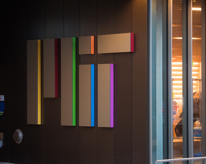The MIT logo placed as panels on the side of a building, with each panel highlighted in different rainbow colors.