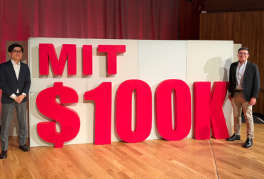 Hiro Ogasawara and Rowell Castro stand to either side of a large sign reading "MIT $100K".