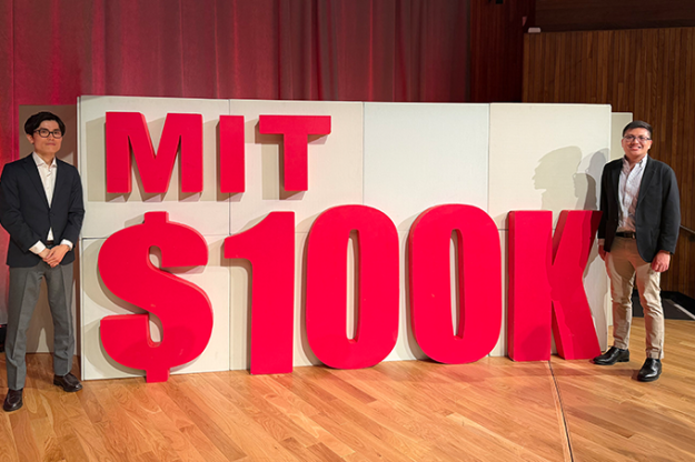 Hiro Ogasawara and Rowell Castro stand to either side of a large sign reading "MIT $100K".