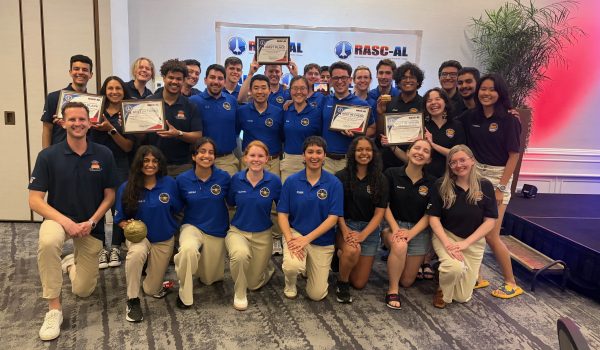A large group of students in blue and navy polo shirts, holding up several award certificates.