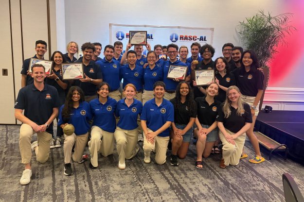 A large group of students in blue and navy polo shirts, holding up several award certificates.