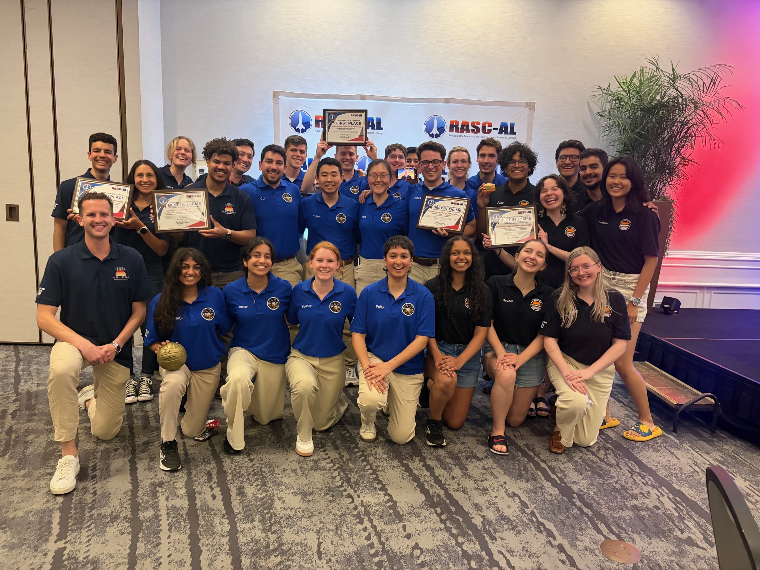 A large group of students in blue and navy polo shirts, holding up several award certificates.