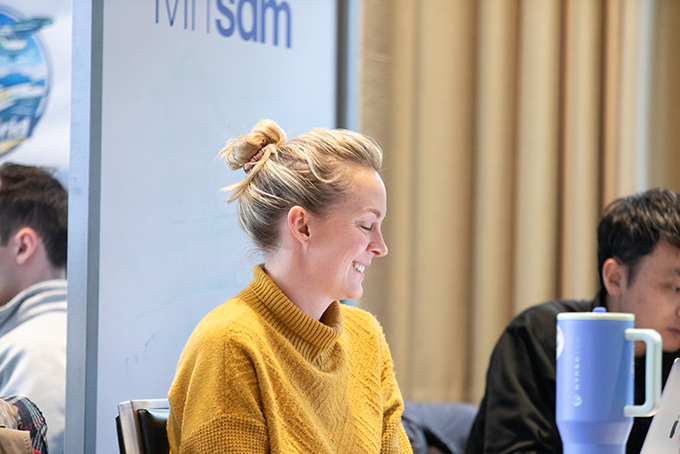 A woman in a yellow sweater sitting in a classroom laughs.